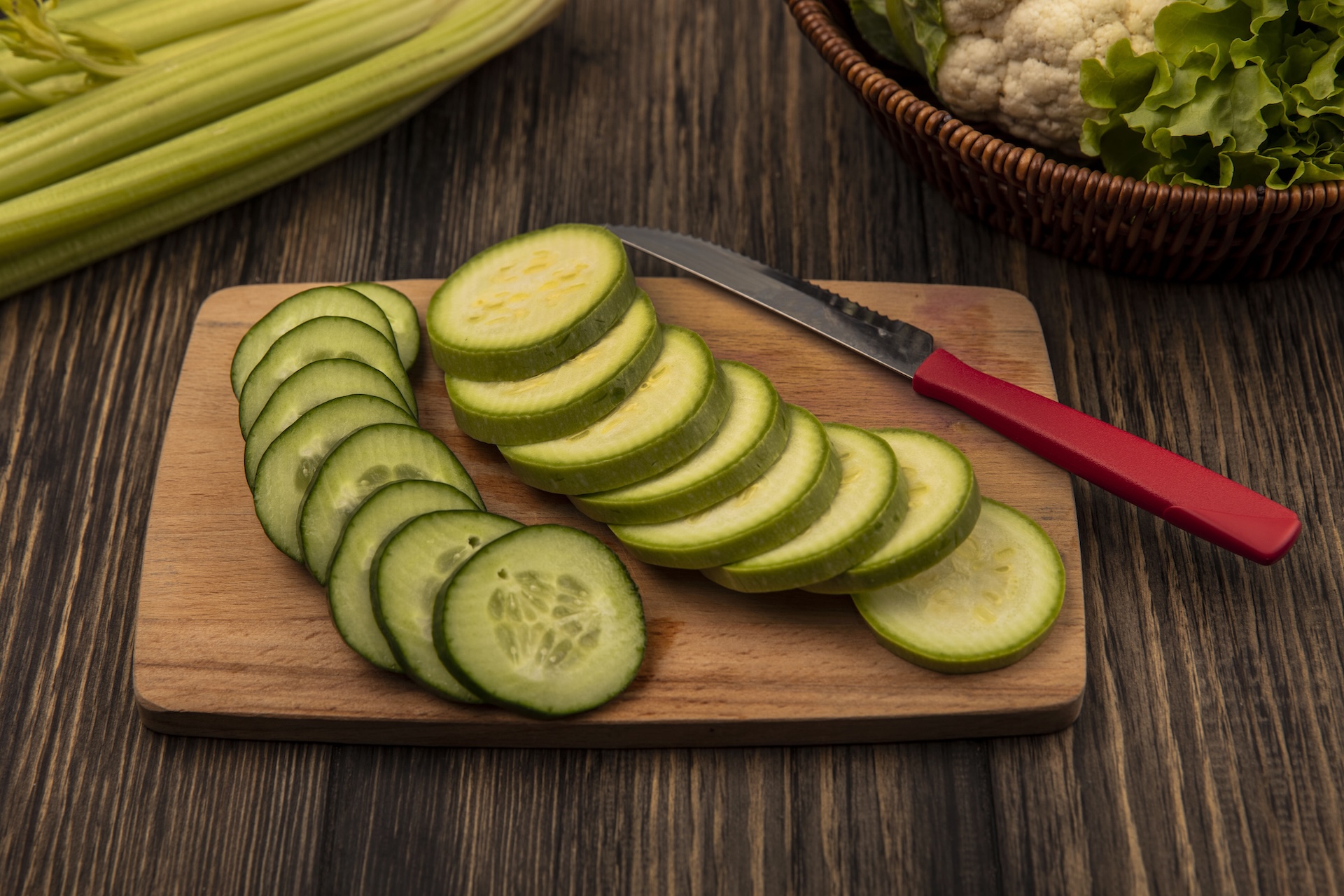 top view of chopped vegetables such as cucumber and zucchinis on a wooden kitchen board with with knife with cauliflower and lettuce on a bucket with celery isolated on a wooden background